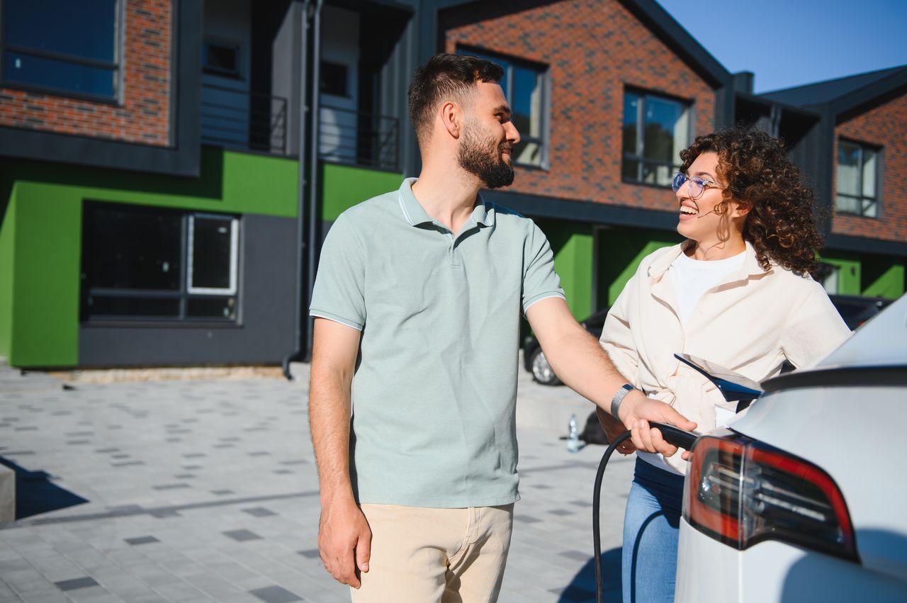 Couple souriant rechargeant leur voiture électrique devant un logement familial, mode de vie sobre et durable.