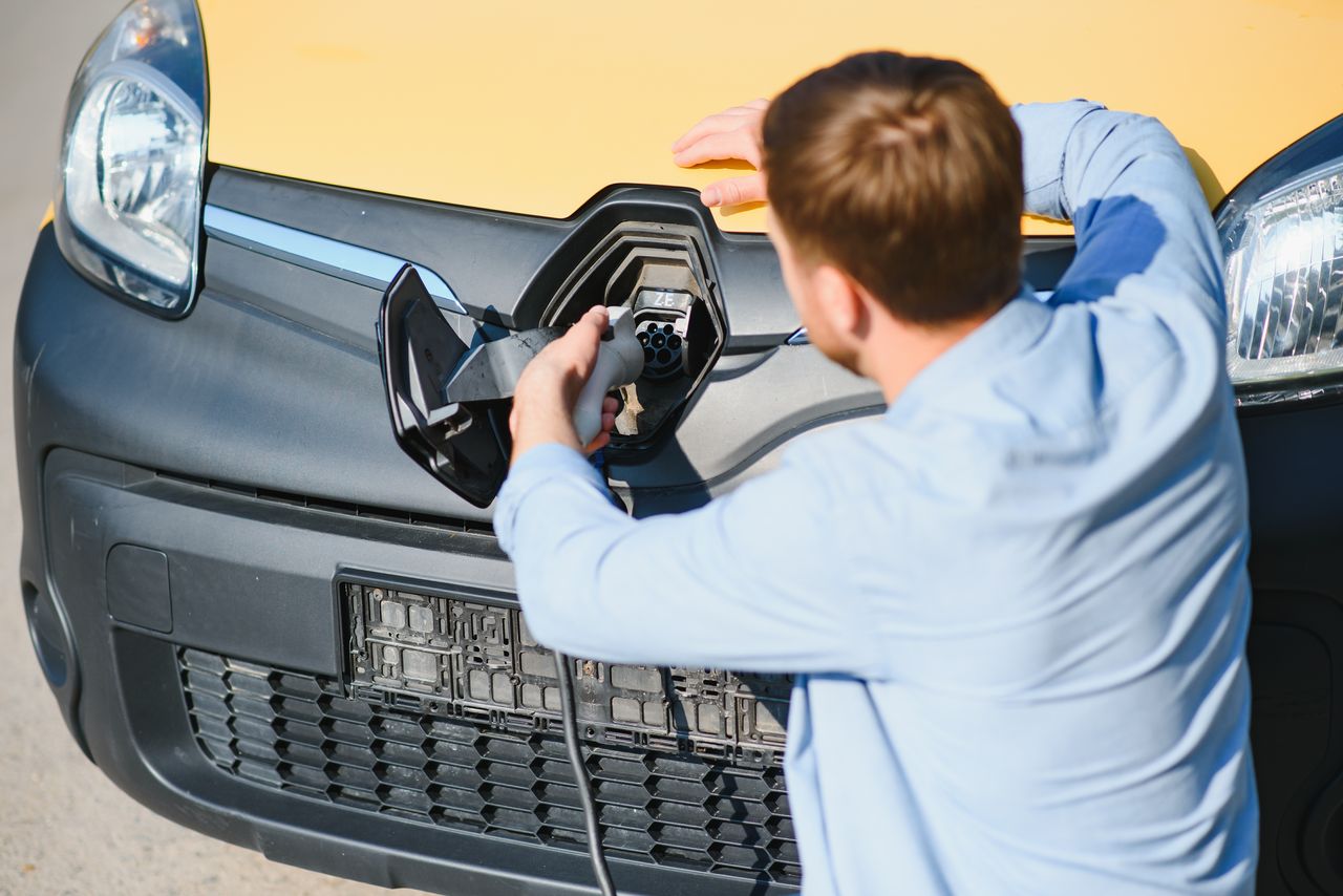 Homme tenant câble devant sa nouvelle voiture électrique, station de recharge et dossier administratif à l'appui.
