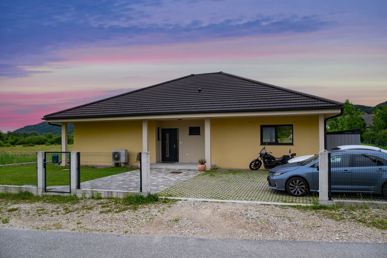 Maison moderne avec place de parking et borne murale alimentant une voiture électrique, façade minimaliste écoénergétique.