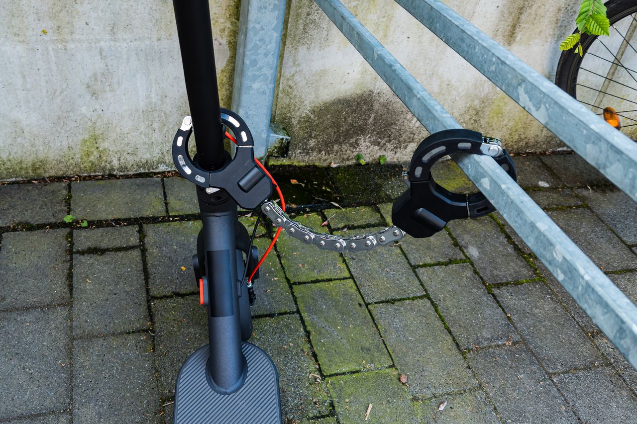 Trottinette électrique verrouillée avec cadenas et casque devant immeuble urbain, sécurité et mobilité partagée en ville.